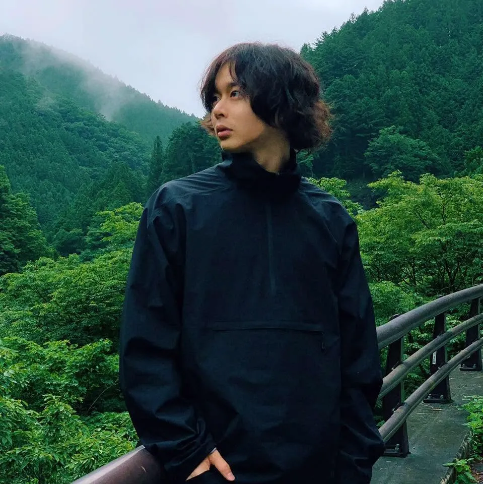 柏木太郎 — 黒いジャケットを着て、緑の山並みを背景に佇むポートレート